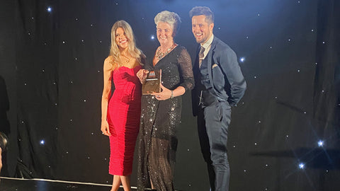 Photo showing Maxine Laceby, a smiling middle aged white woman with short grey hair, standing in between a smiling white man and smiling white woman on a stage. She is holding an award and wearing a glamorous black outfit.