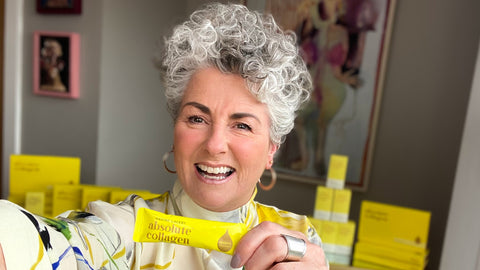 Photo showing Maxine Laceby, a smiling white woman with short grey curly hair, holding up a sachet of Absolute Collagen while taking a selfie and smiling