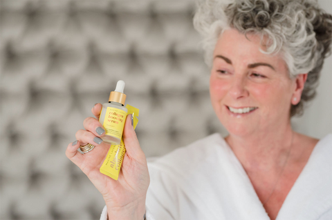 Maxine Laceby holding a yellow Absolute Collagen serum bottle and sachet and smiling against a grey background