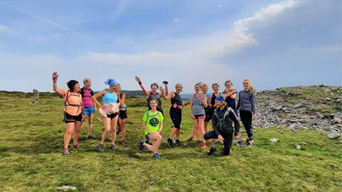Photo of a group of white woman in colourful running gear standing on a hill with the blue sky in the background and a white man crouching in front of them