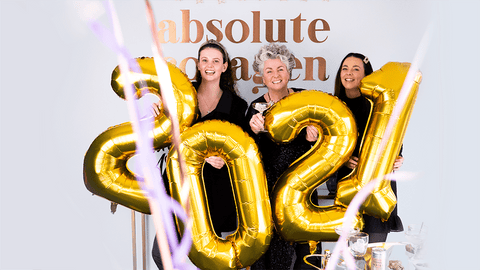 Photo showing Maxine, Margot and Darcy Laceby smiling in front of an Absolute Collagen sign and holding large gold balloons in a 2021 shape while confetti falls around them