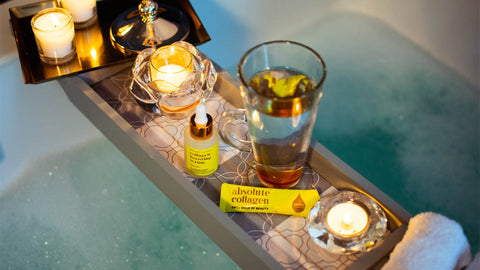 Photo showing a relaxing bubble bath with wooden bath tray which contains a latte glass, candles, flannels, Absolute Collagen sachet and Maxerum