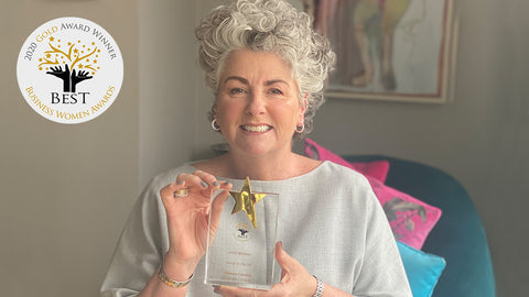 Photo showing Maxine Laceby sitting in front of some framed art and holding up a Best Business Woman awards trophy, she is wearing a grey top and smiling, and the Best Business Woman awards logo is visible in the top left