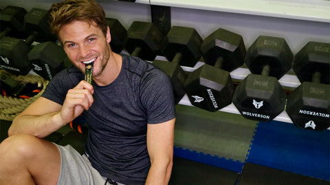 Photo of a white man with brown hair smiling and taking an Absolute Collagen sachet with weight lifting equipment in the background
