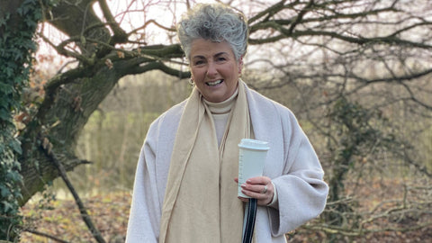 Photo showing Maxine Laceby standing outside and smiling, she is holding a reusable Starbucks coffee cup, wearing a long cream coat, and has her lurcher Rosie standing beside her