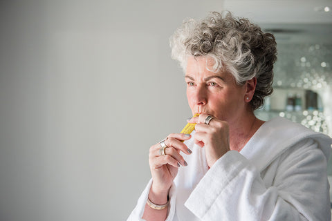 Photo of a white woman with short silver hair wearing a white dressing gown and drinking from a yellow Absolute Collagens sachet