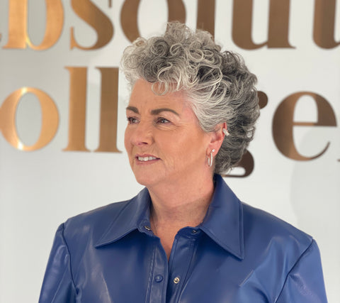 Photo showing a white woman with short wavy silver hair smiling and glancing to the left, she is wearing a blue leather top and standing against the words Absolute Collagen in metallic lettering